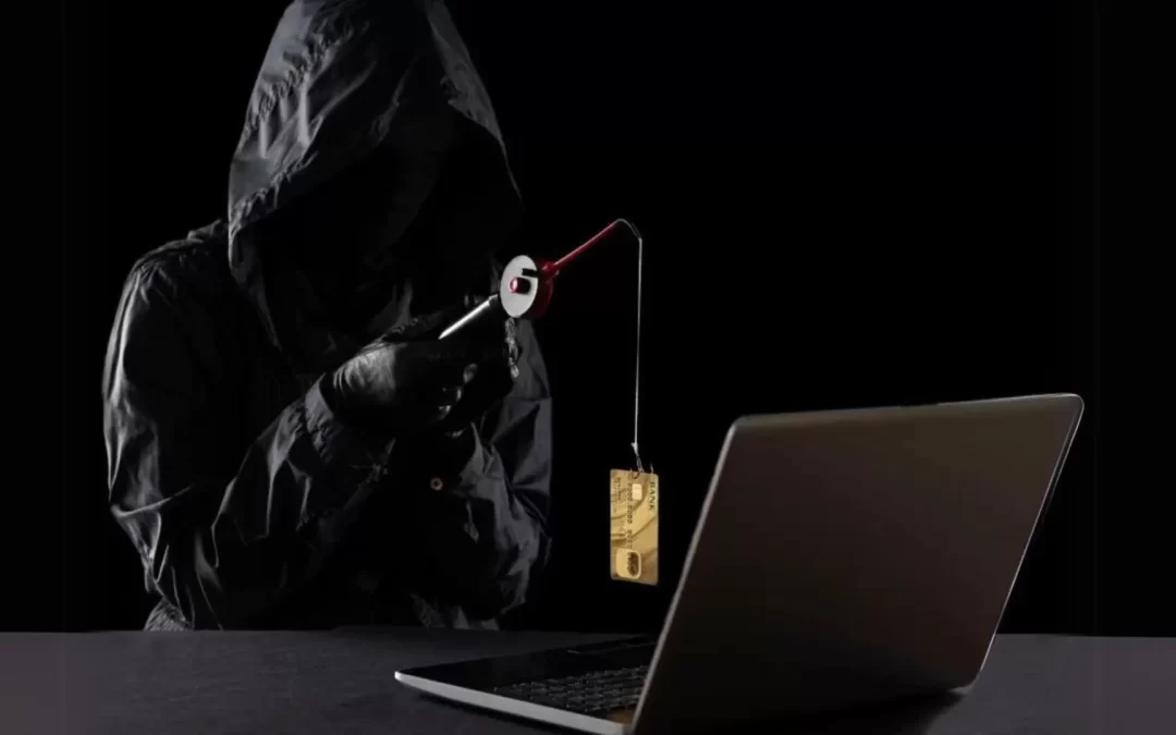 A person dressed in all black, including a jacket and gloves, holding a fishing hook with a credit card on it, positioned over a laptop, hinting at an act of cyber fraud or phishing.