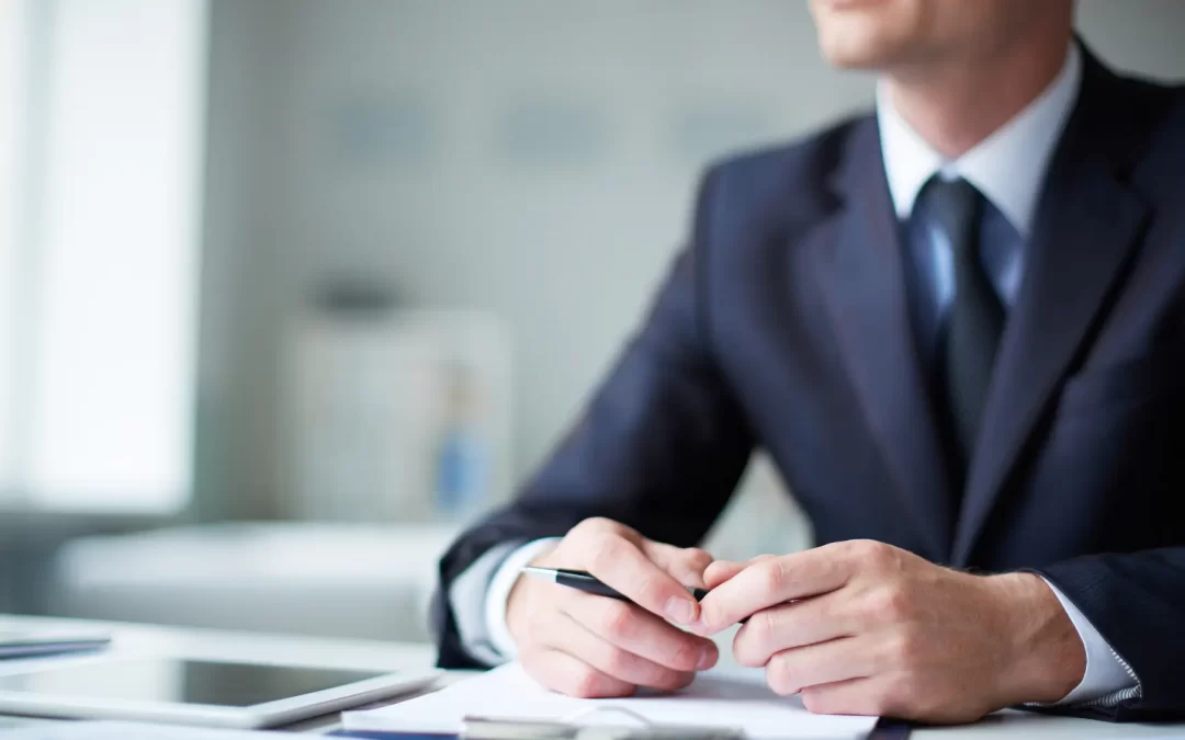 A CEO seated at his desk in his office, with his hands resting on the desk and holding a pen.