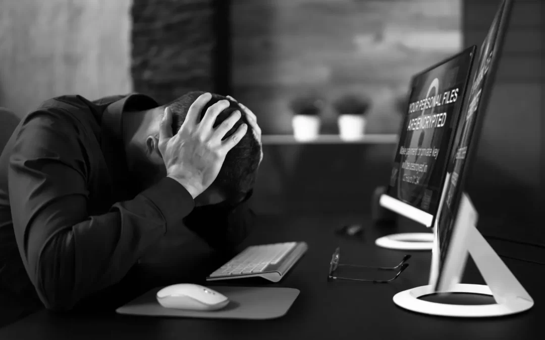 A person sitting at a desk, holding their head in distress, while looking at a computer screen that displays a ransomware message: "YOUR PERSONAL FILES ARE ENCRYPTED." The image conveys a sense of frustration and urgency, highlighting the impact of a cybersecurity breach.